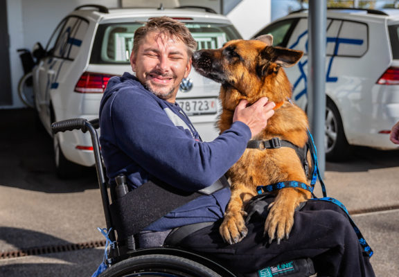 Sozialhund schnuppert unter Aufsicht an Gesicht eines Rollstuhlfahrers, der ihn liebkost