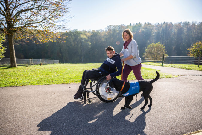 Halterin Sozialhund schiebt Rollstuhlfahrer während ihr Hund an der Leine mitläuft